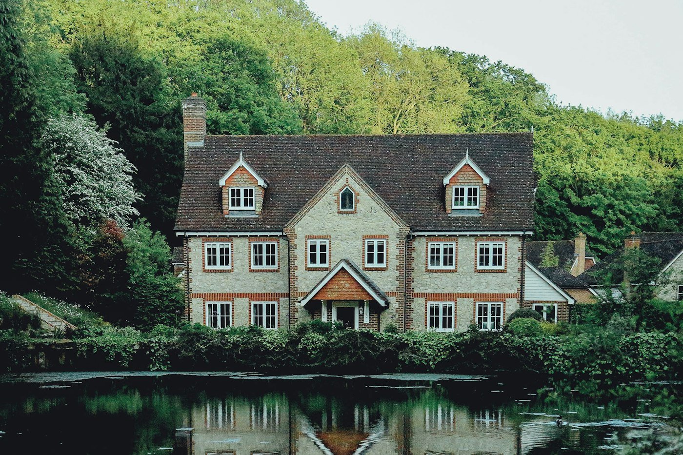 A stone and brick house with a tiled roof, white windows, and trees behind it