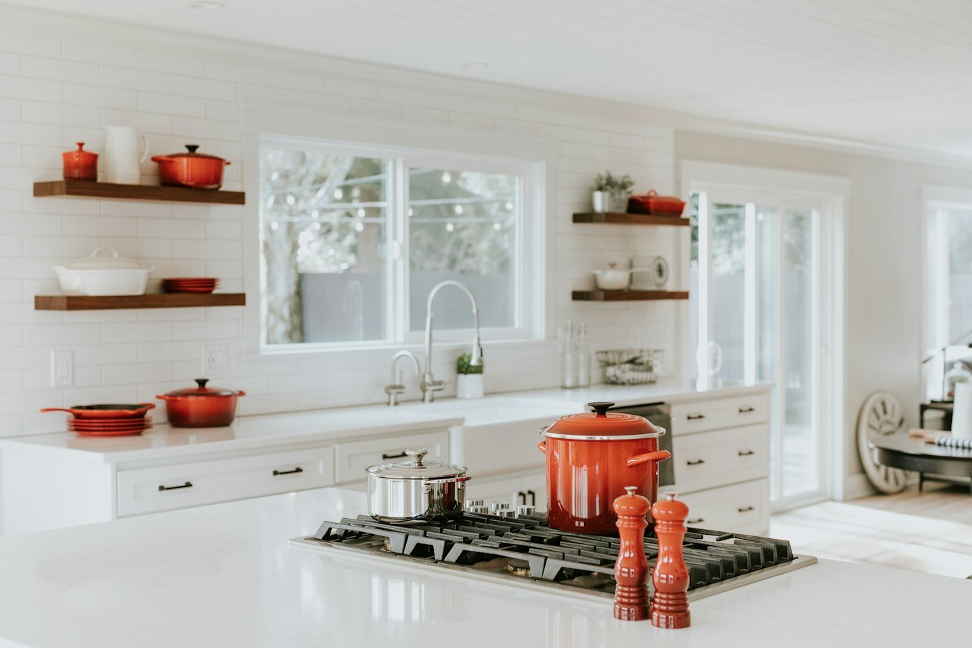 White kitchen with a farmhouse sink, open shelves, and red pots on the island hob